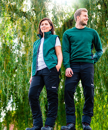 Zwei Personen in grner Gartenarbeitskleidung stehen nebeneinander vor einem Hintergrund mit herabhngenden sten.