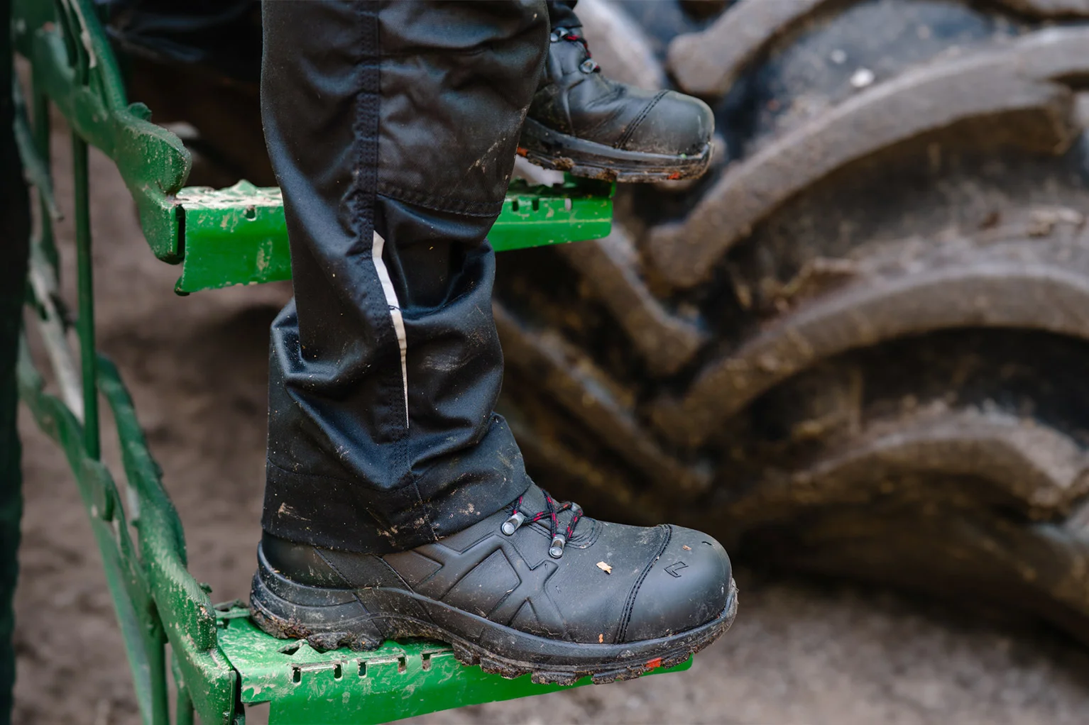 Nahaufnahme von schwarzen Arbeitsschuhen mit roter Schn�rung auf einer gr�nen Metallstufe; im Hintergrund ist ein gro�er, stark verschmutzter Maschinenreifen zu sehen.