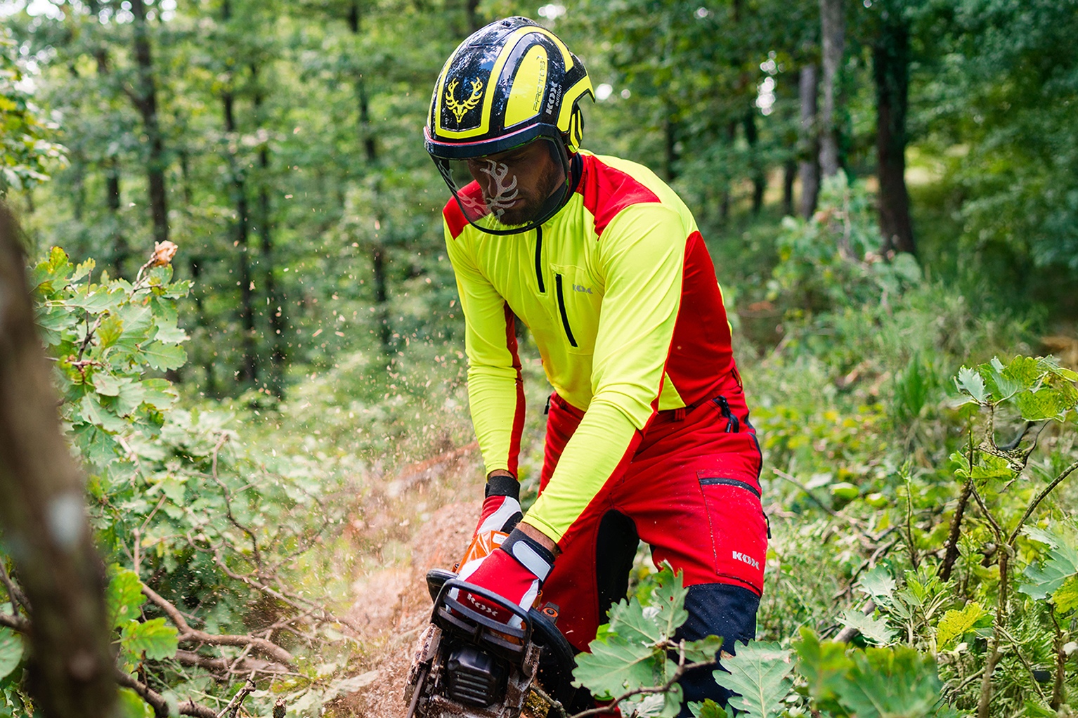 Forstarbeiter mit Motorsge bei der Arbeit im Wald, getragen wird ein KOX Funktionsshirt in leuchtendem Orange mit schwarzen Einstzen sowie ein Schutzhelm mit Visier und Gehrschutz.