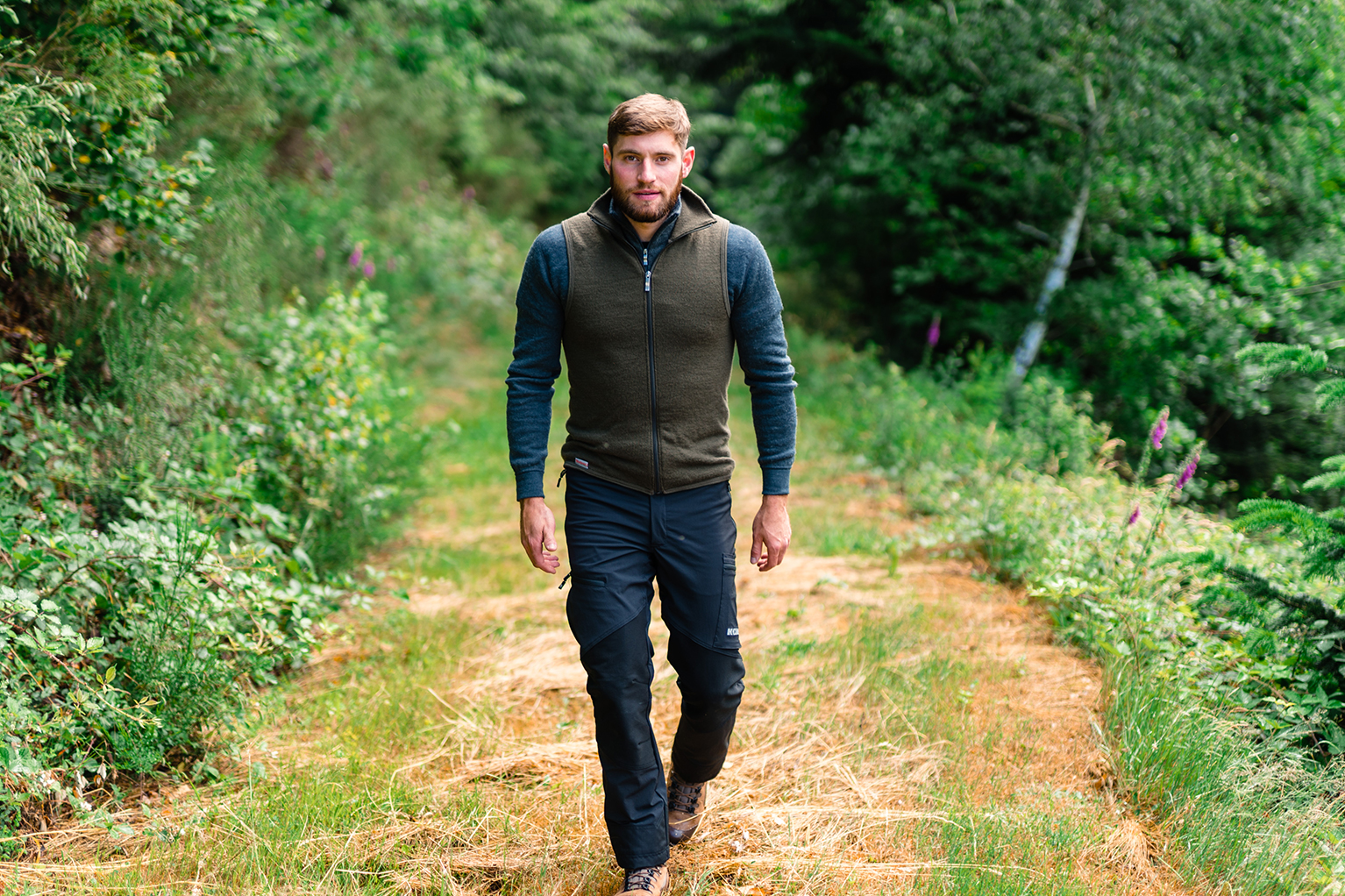 Ein Mann geht auf einem Waldweg, umgeben von grnem Wald. Er trgt eine Fleecejacke, Outdoor-Hose und Wanderschuhe.