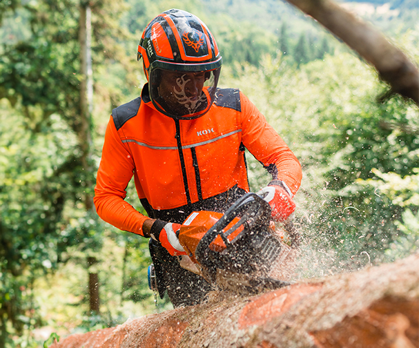Person in leuchtend orangefarbener Forstschutzkleidung sgt mit Motorsge liegenden Baumstamm; Holzspne fliegen, dichter Wald im Hintergrund.