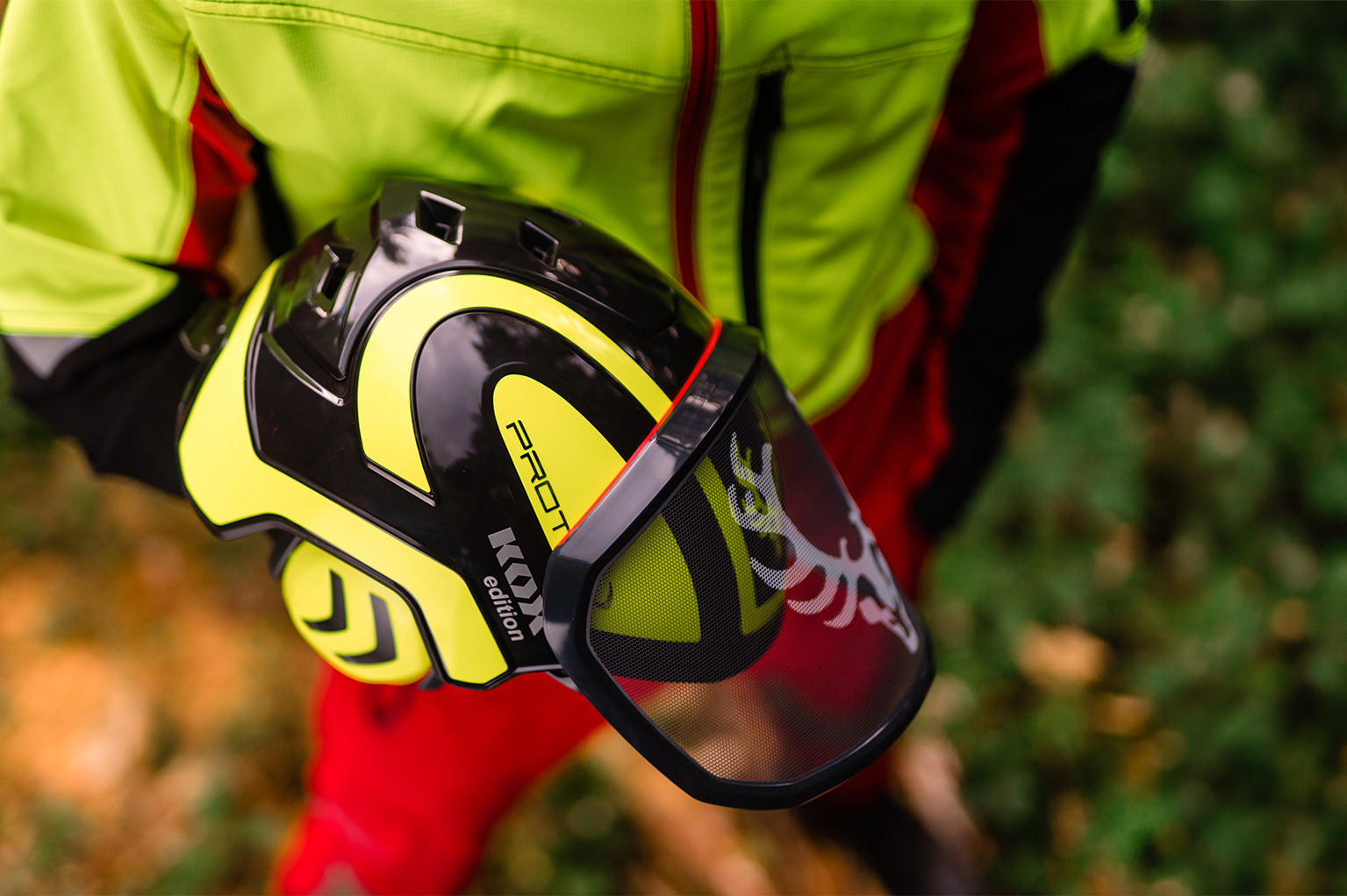 Eine Person in gelb-roter Schutzkleidung h�lt einen gelb-schwarzen Forsthelm mit Visier im Wald.